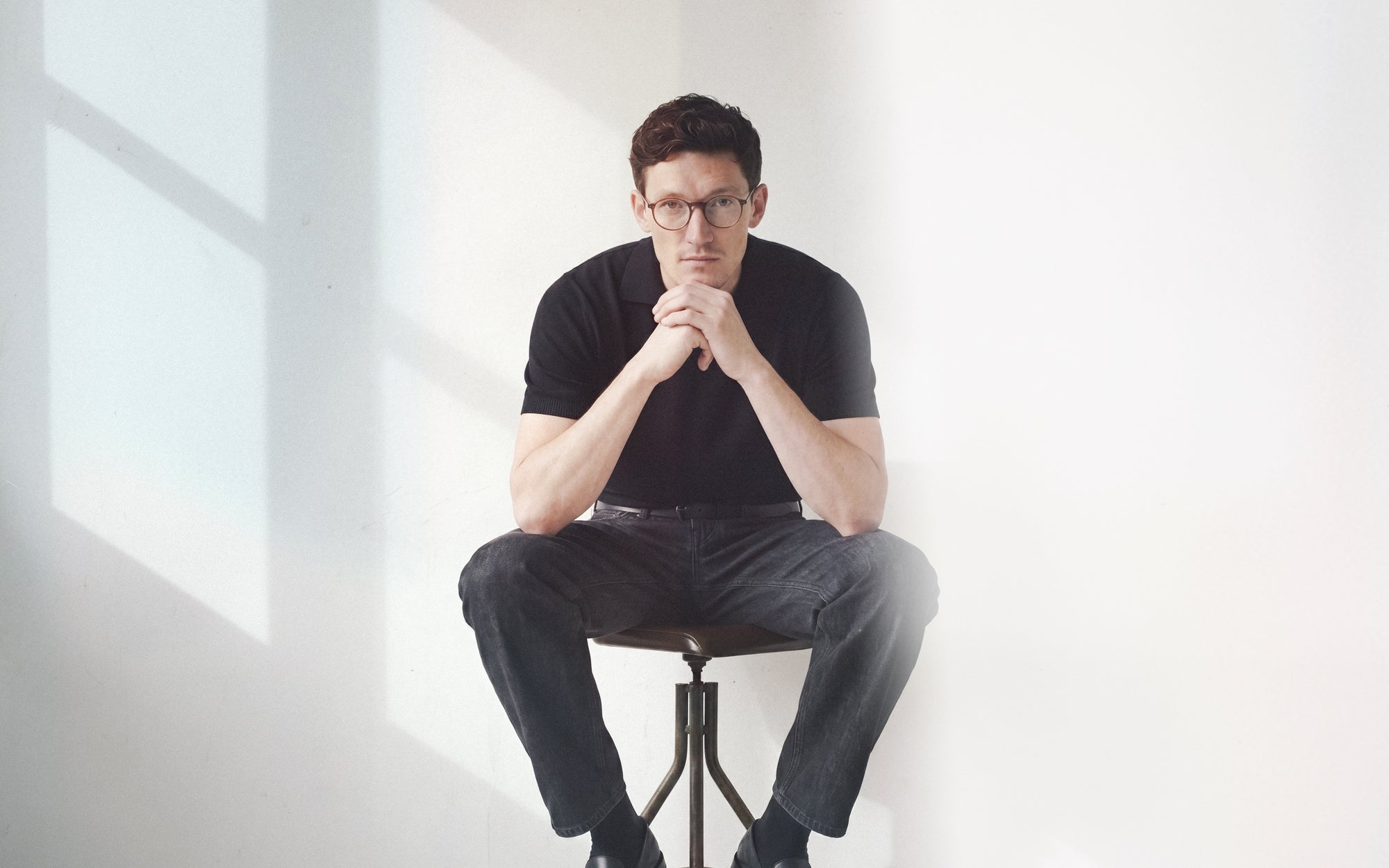 Editorial portrait of man with glasses in black polo sitting on stool in soft window light