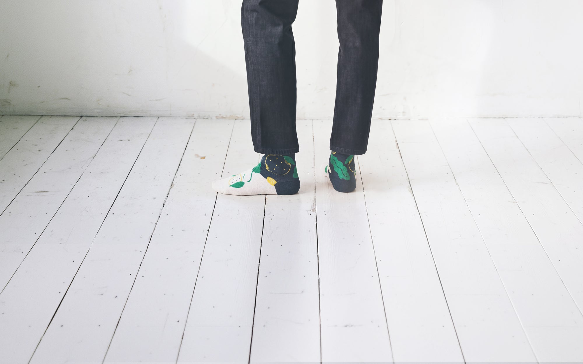 Studio photo of man in dark jeans on a white wooden floor, wearing Limono Lemonheads Black socks