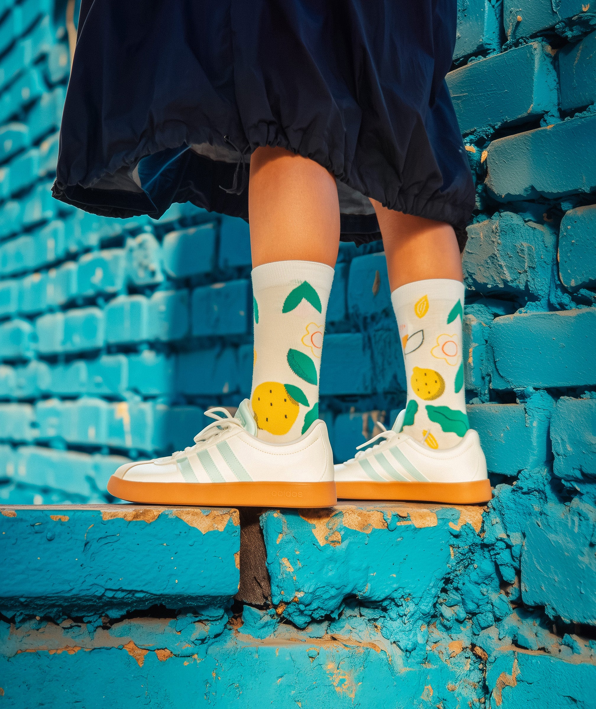 Person wearing colorful Limono Lemonheads socks and white Adidas sneakers against a blue brick wall.