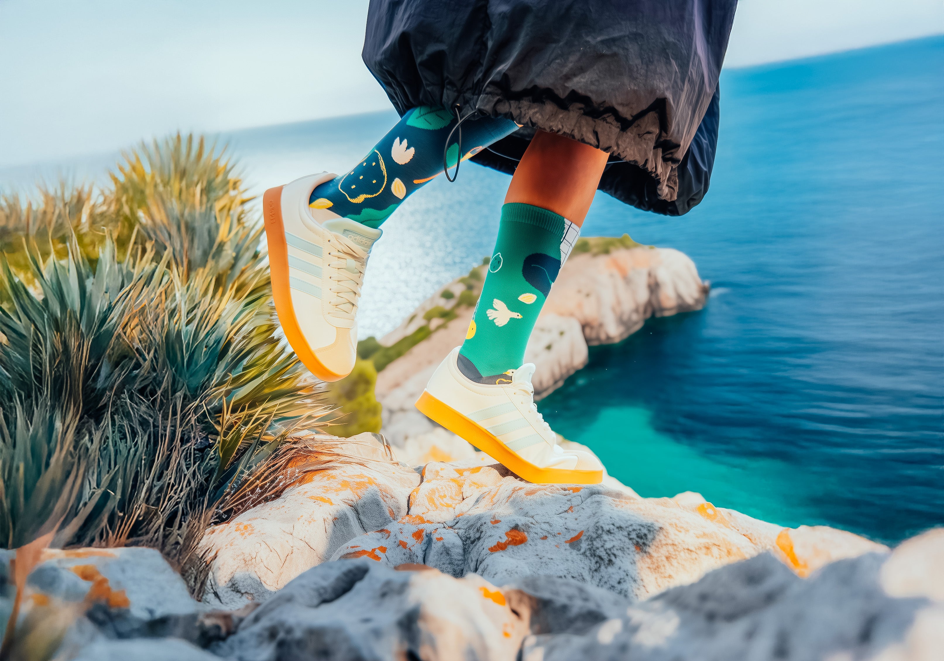 Person wearing Limono Lemonheads socks with white patterns and white sneakers standing on a rocky edge overlooking water.