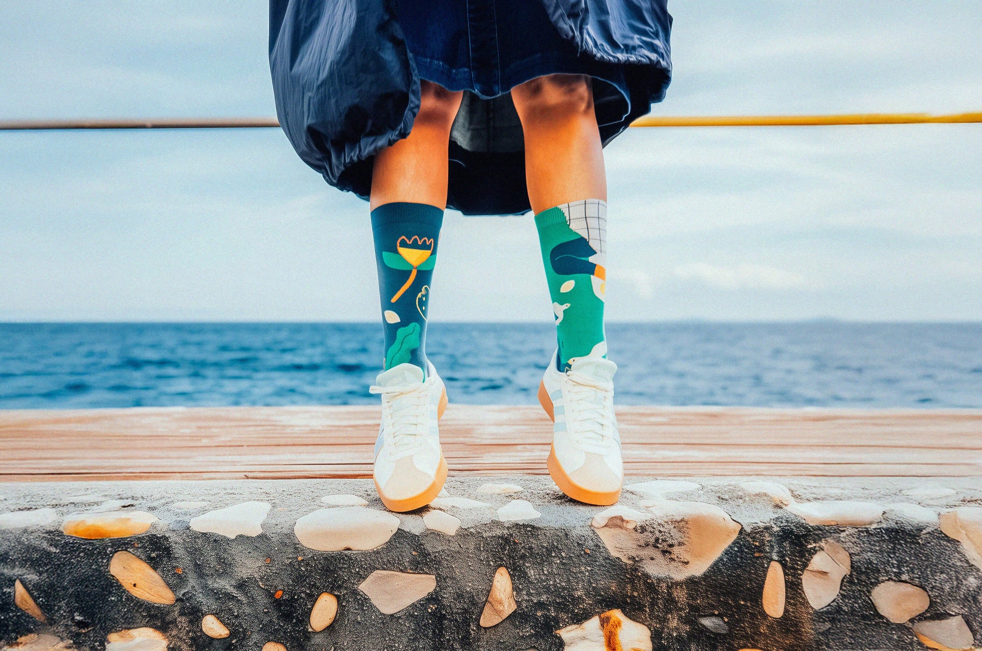 Person wearing colorful Limono Lemonheads socks and white sneakers standing on a ledge with ocean view