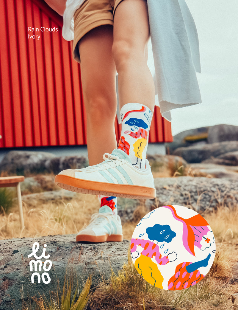 Limono Socks Rain Clouds collection. Illustrated socks on trend made in Portugal. Organic combed cotton. Person wearing colorful socks with a nature-themed design, standing on a rock with a red building in the background.