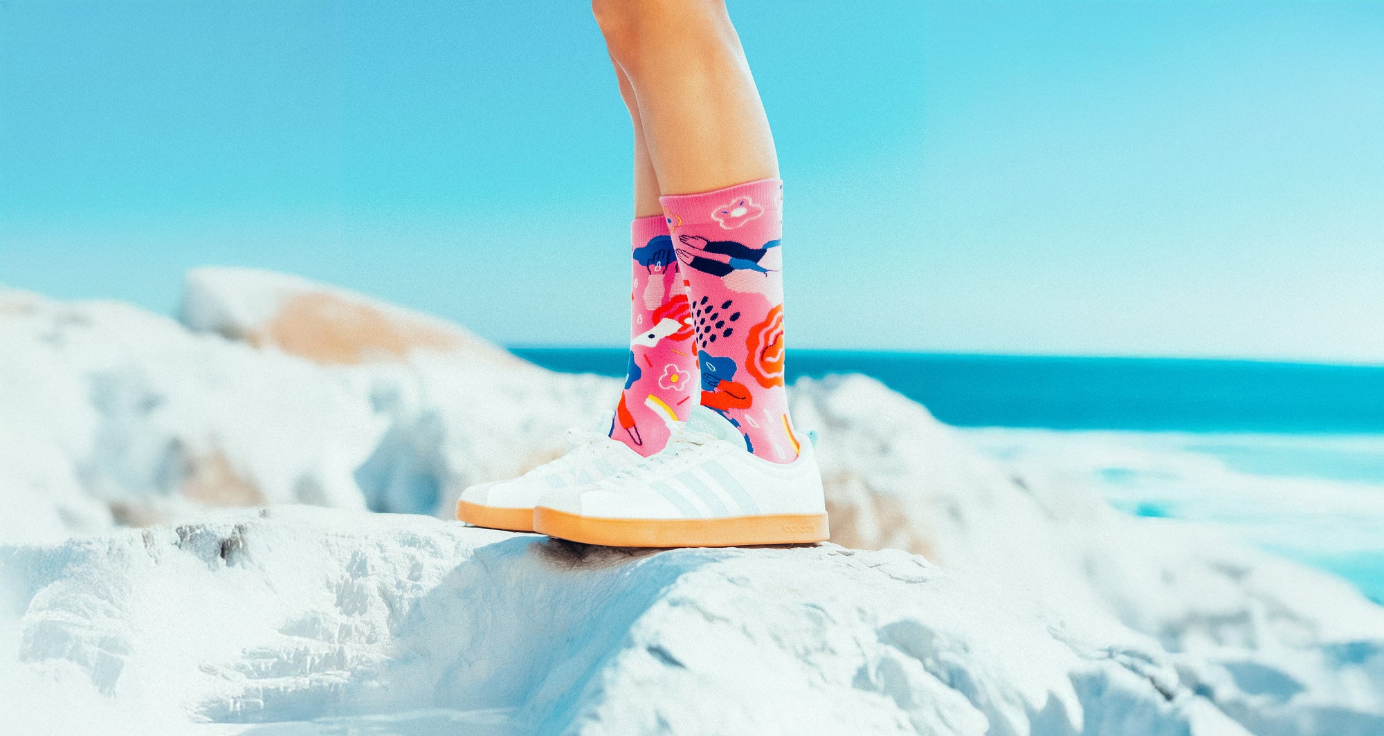 Person wearing colorful Limono Rain Clouds socks and white shoes on a snowy landscape with a blue sky.