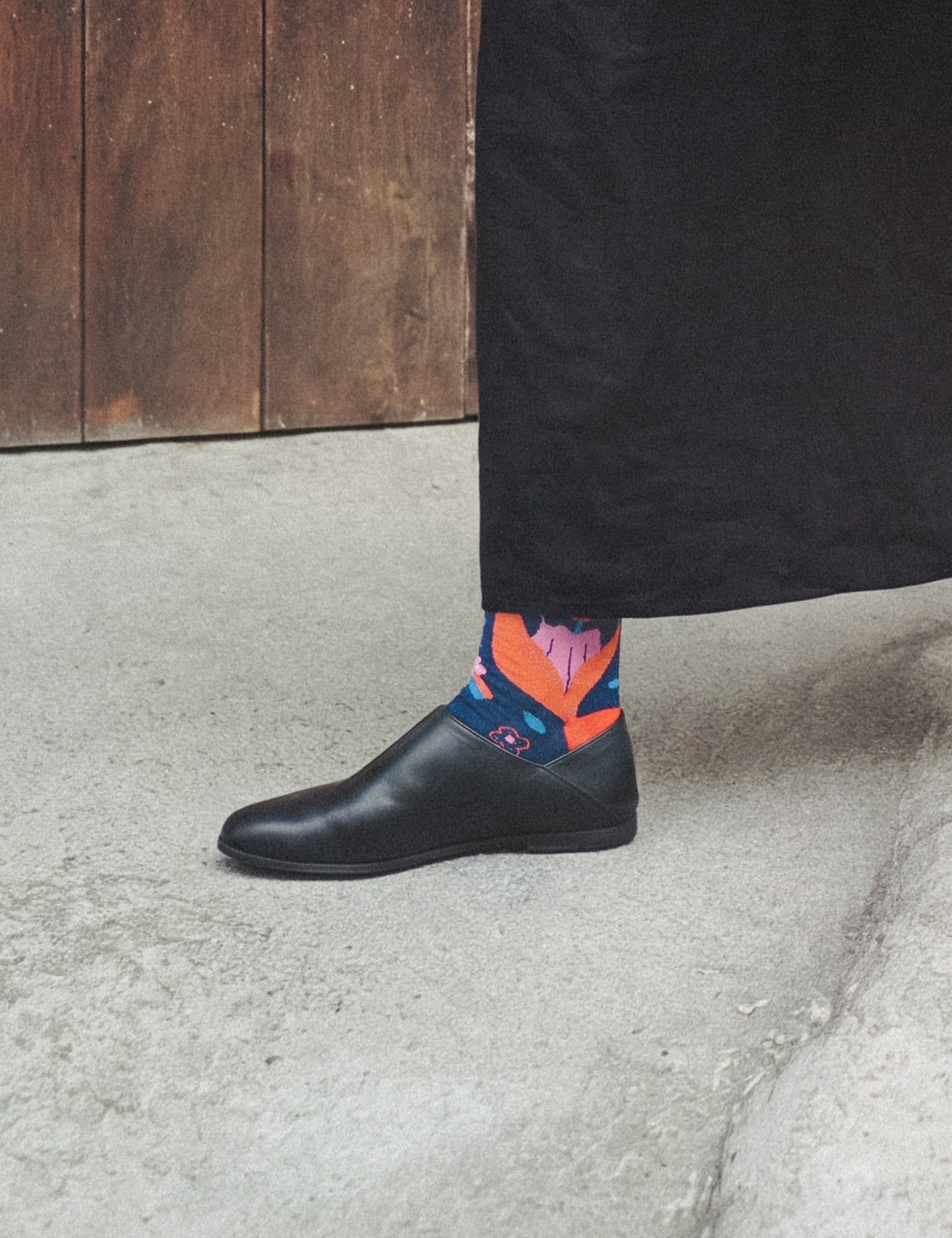 Person in long black skirt and black slipper on concrete by a wooden door, wearing Limono Rain Clouds Black socks
