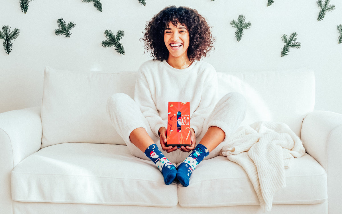 A smiling woman with curly hair sits cross-legged on a white sofa, holding a red box. She is wearing a white sweatshirt and sweatpants, with blue patterned socks visible.