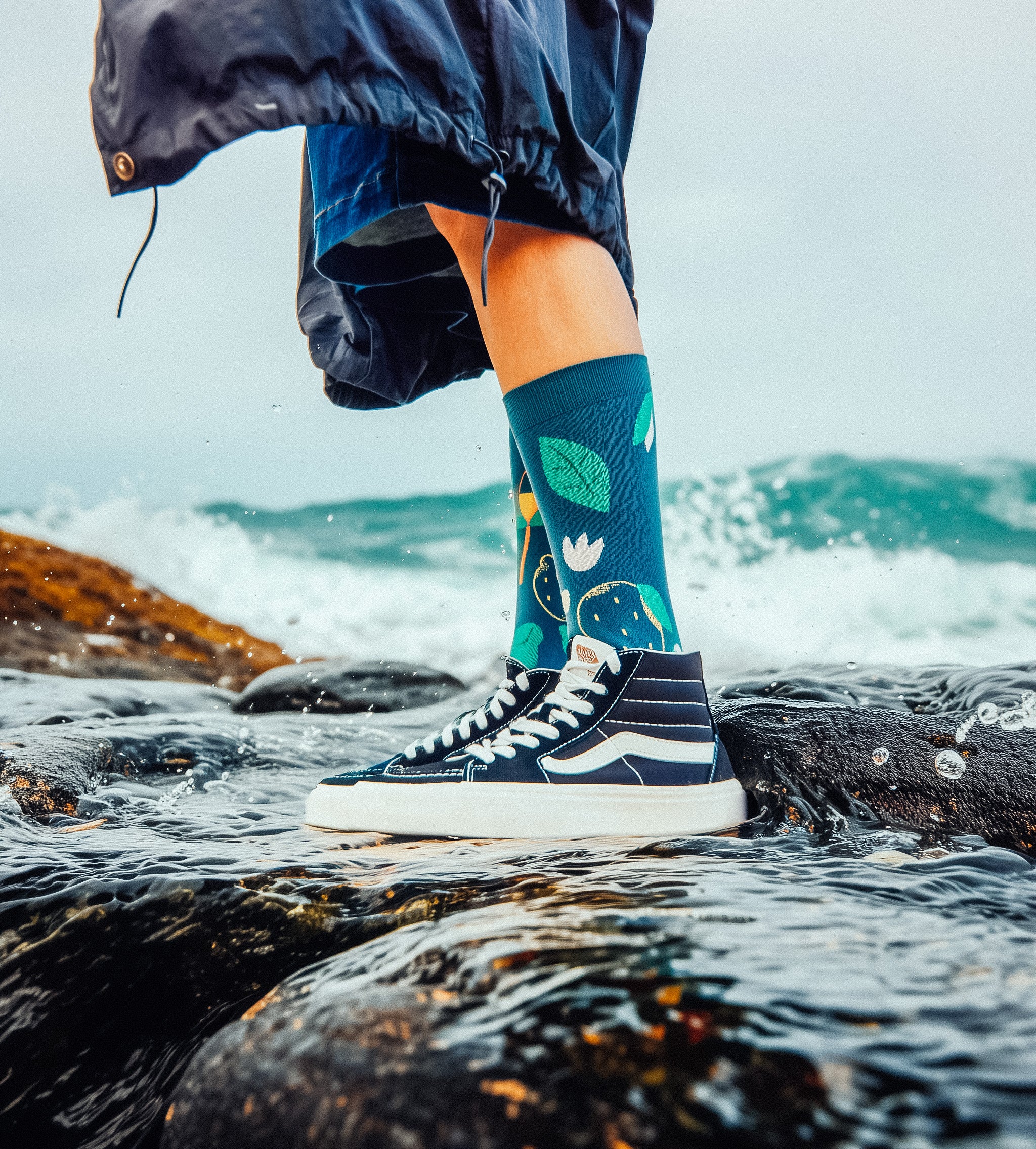 Limono Socks Lemonheads collection. Illustrated socks on trend made in Portugal. Organic combed cotton. Person wearing colorful socks and high-top sneakers on a rocky shoreline with ocean waves.