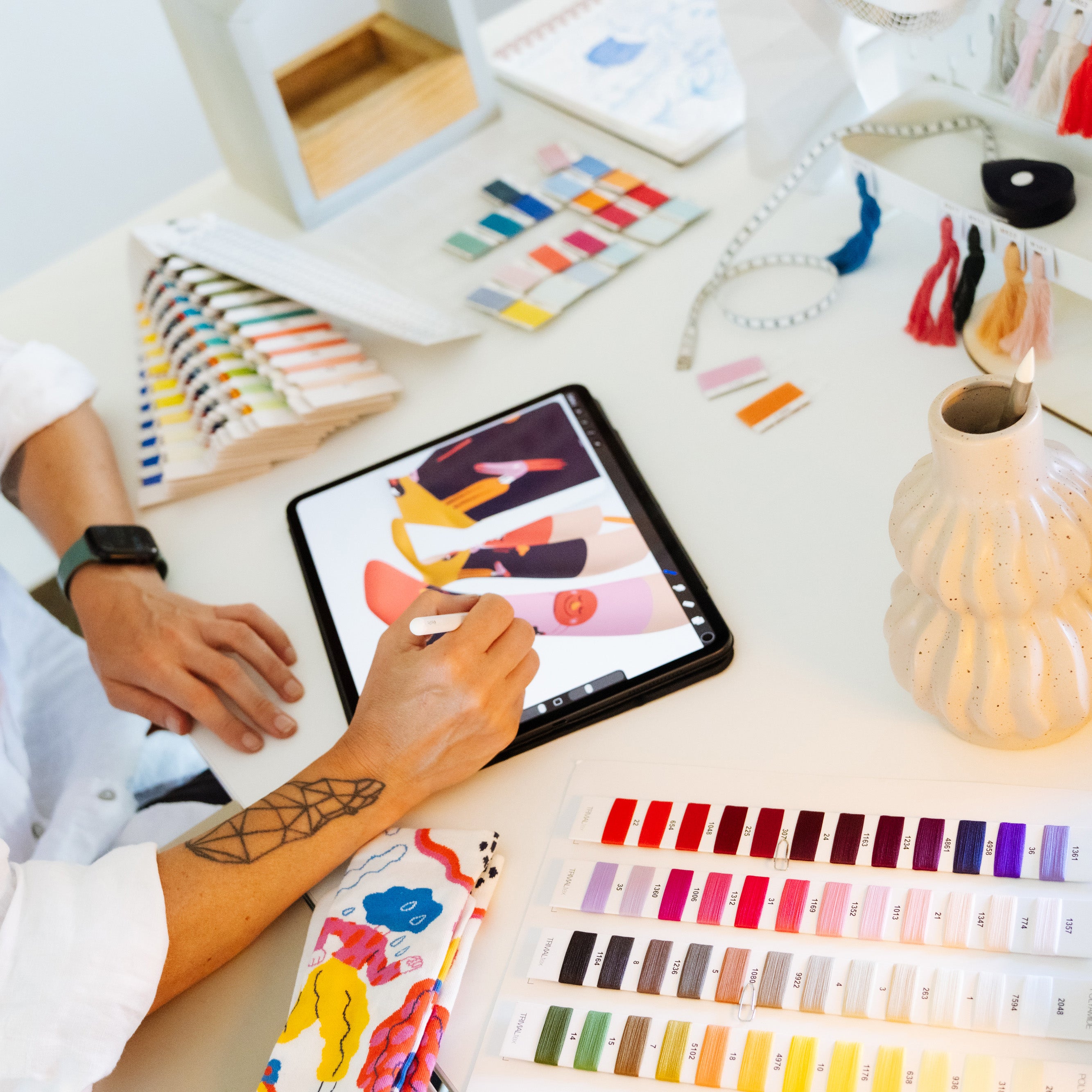 Studio top view of designer hands working on iPad with color thread swatches and ceramic vase