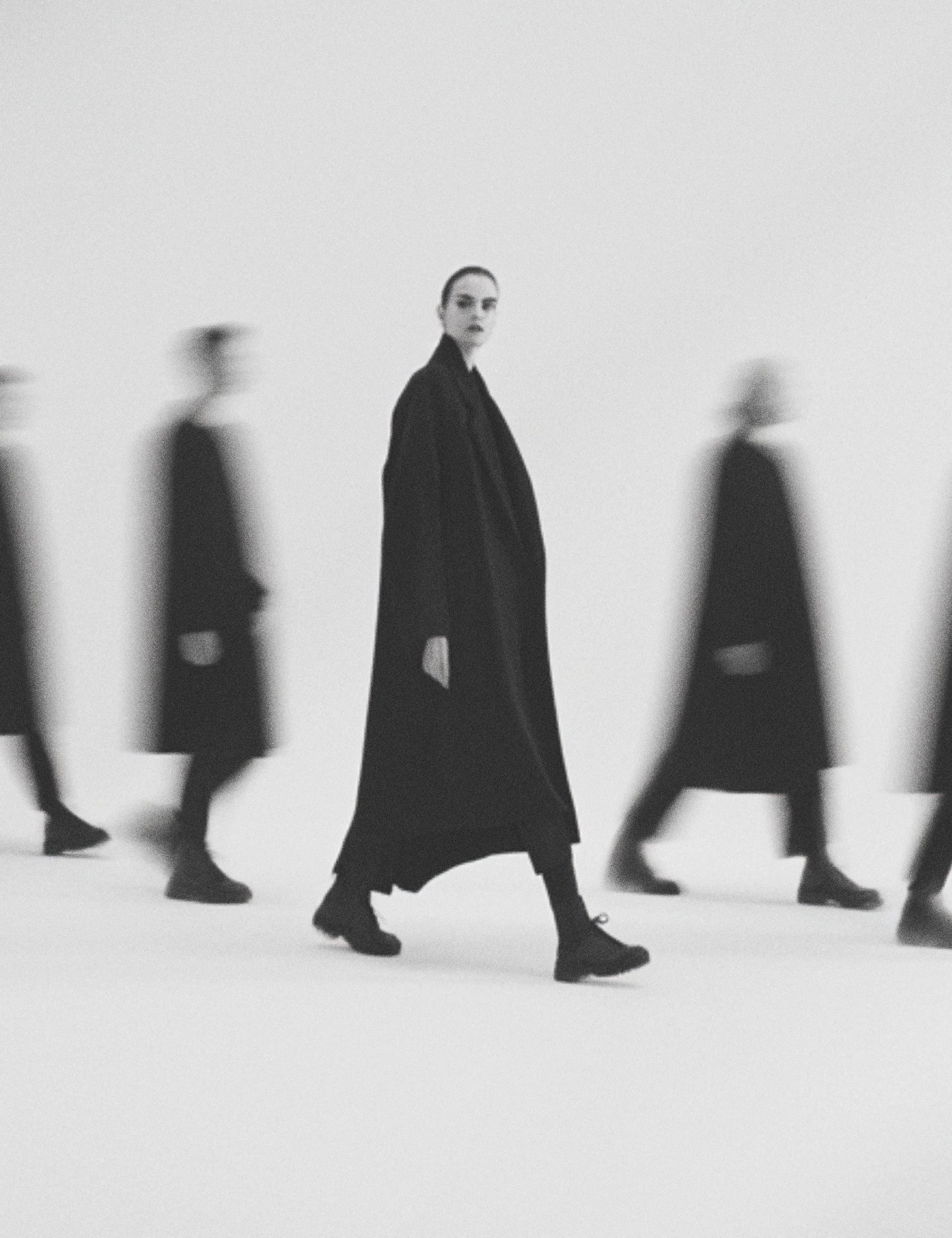 Fashion editorial of woman in long black coat with blurred figures behind, black and white minimalist