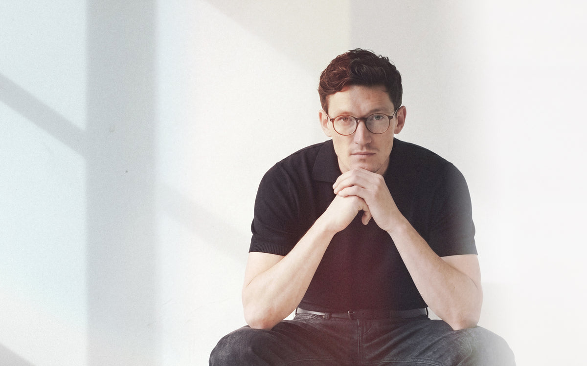 Editorial close portrait of man with glasses in black polo with chin resting on hands