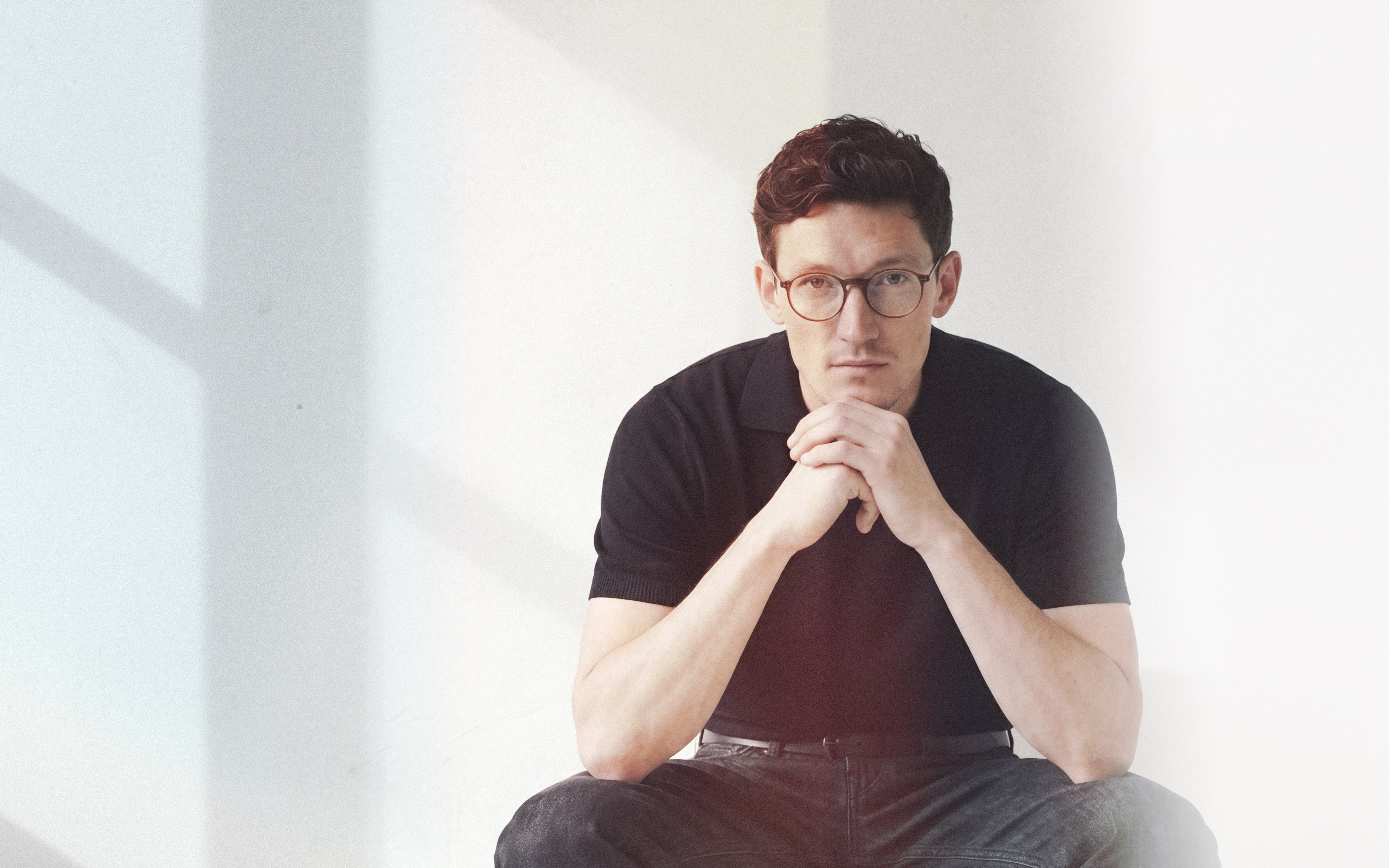 Editorial close portrait of man with glasses in black polo with chin resting on hands