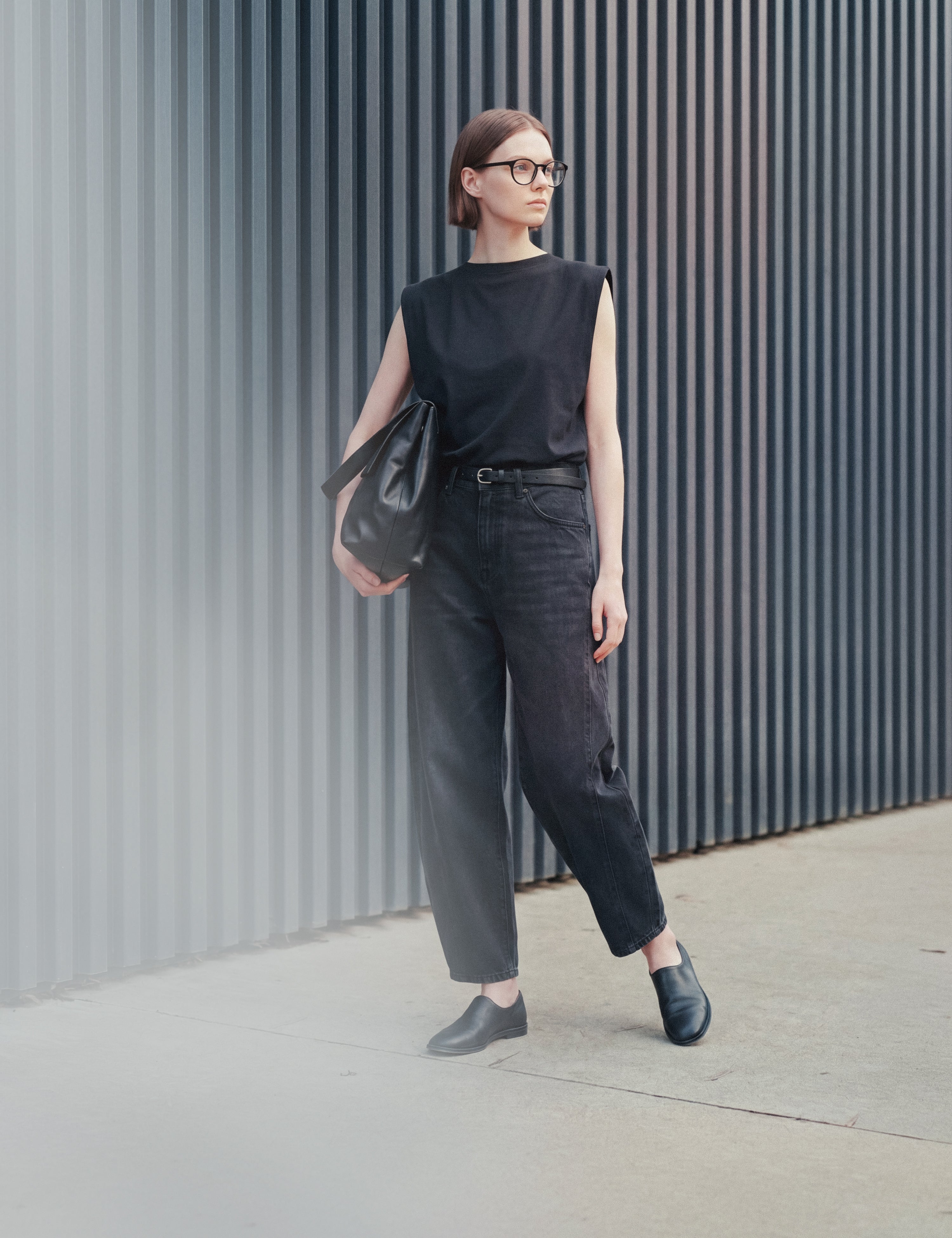 Woman in black sleeveless top and dark jeans with leather bag by ribbed grey wall