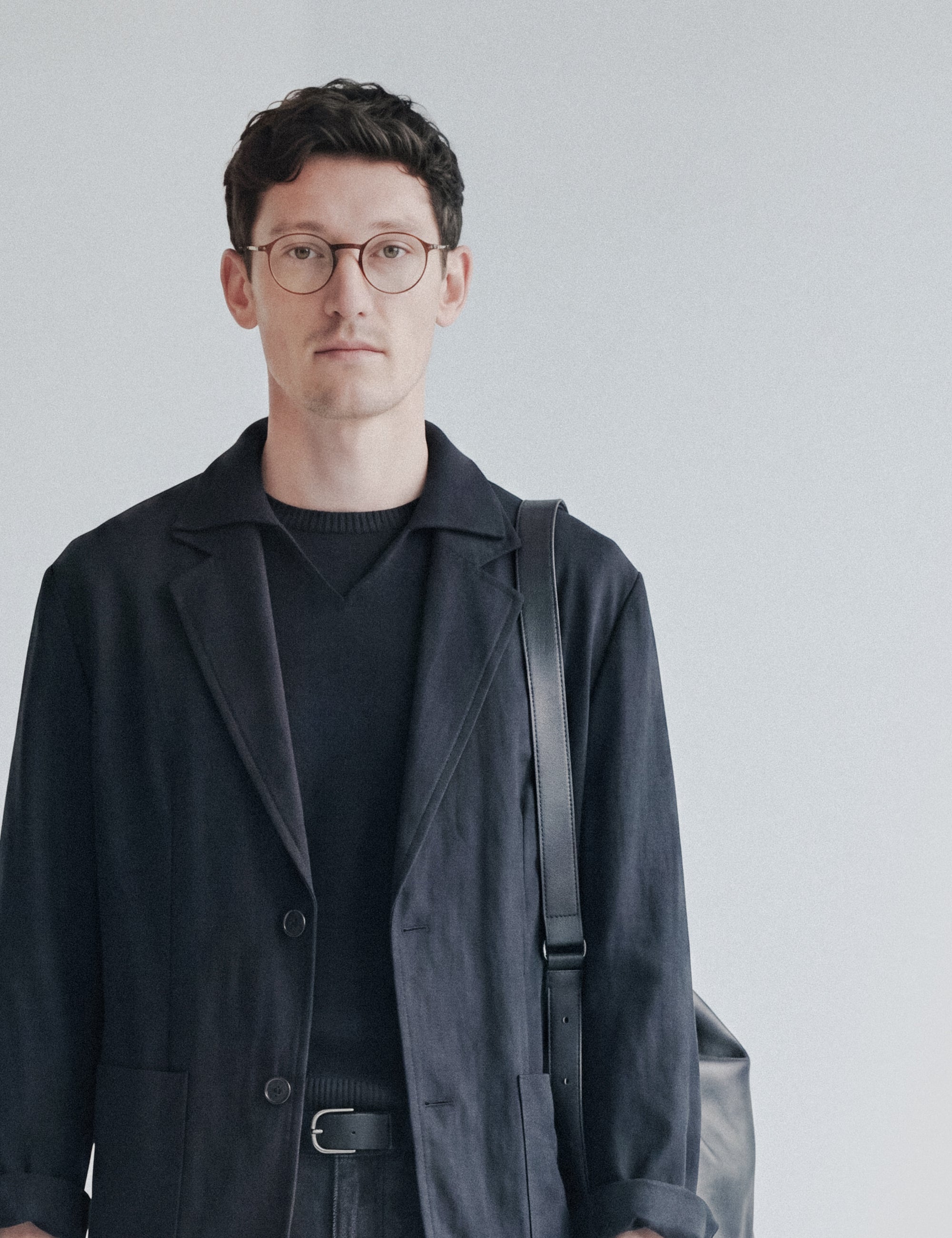 Editorial portrait, man in glasses with dark jacket and leather crossbody bag strap