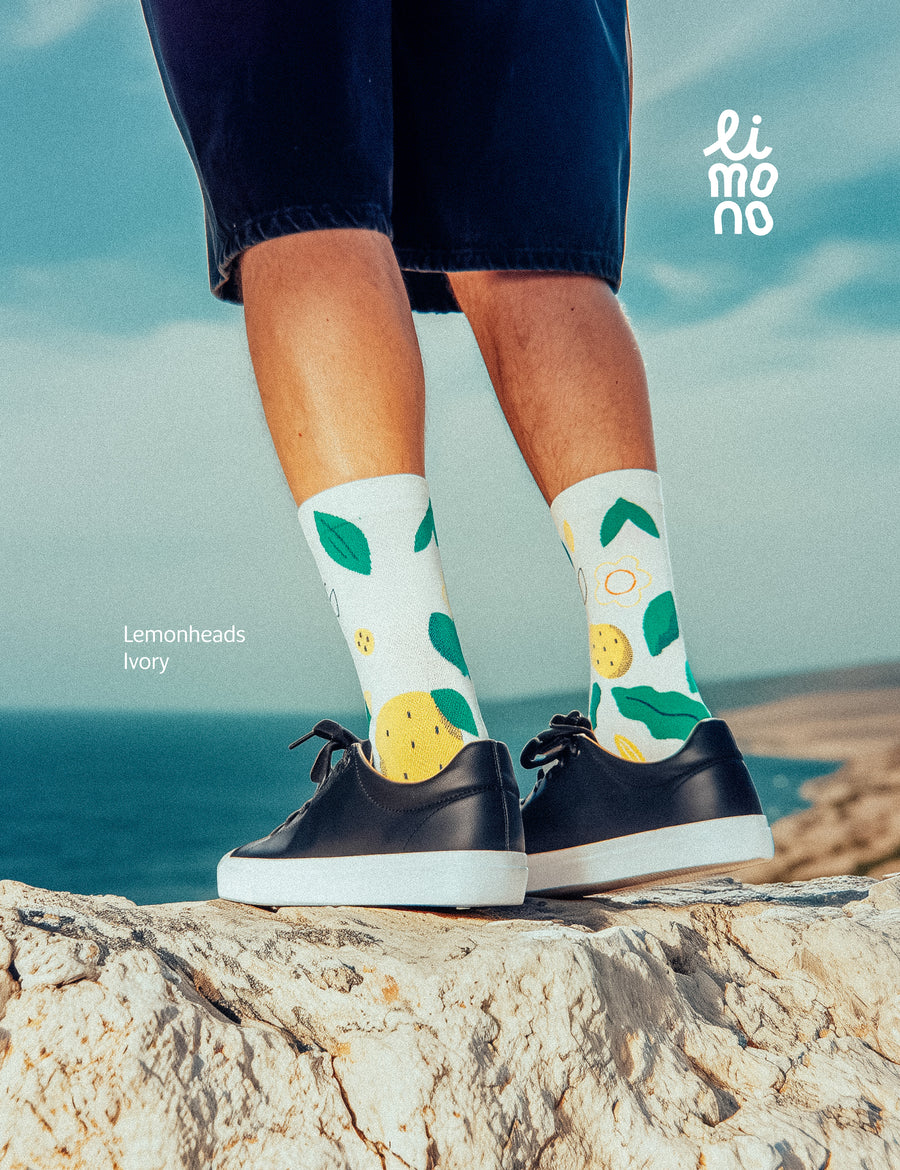 Cropped legs on rock with dark sneakers wearing Limono Lemonheads Ivory socks by the ocean
