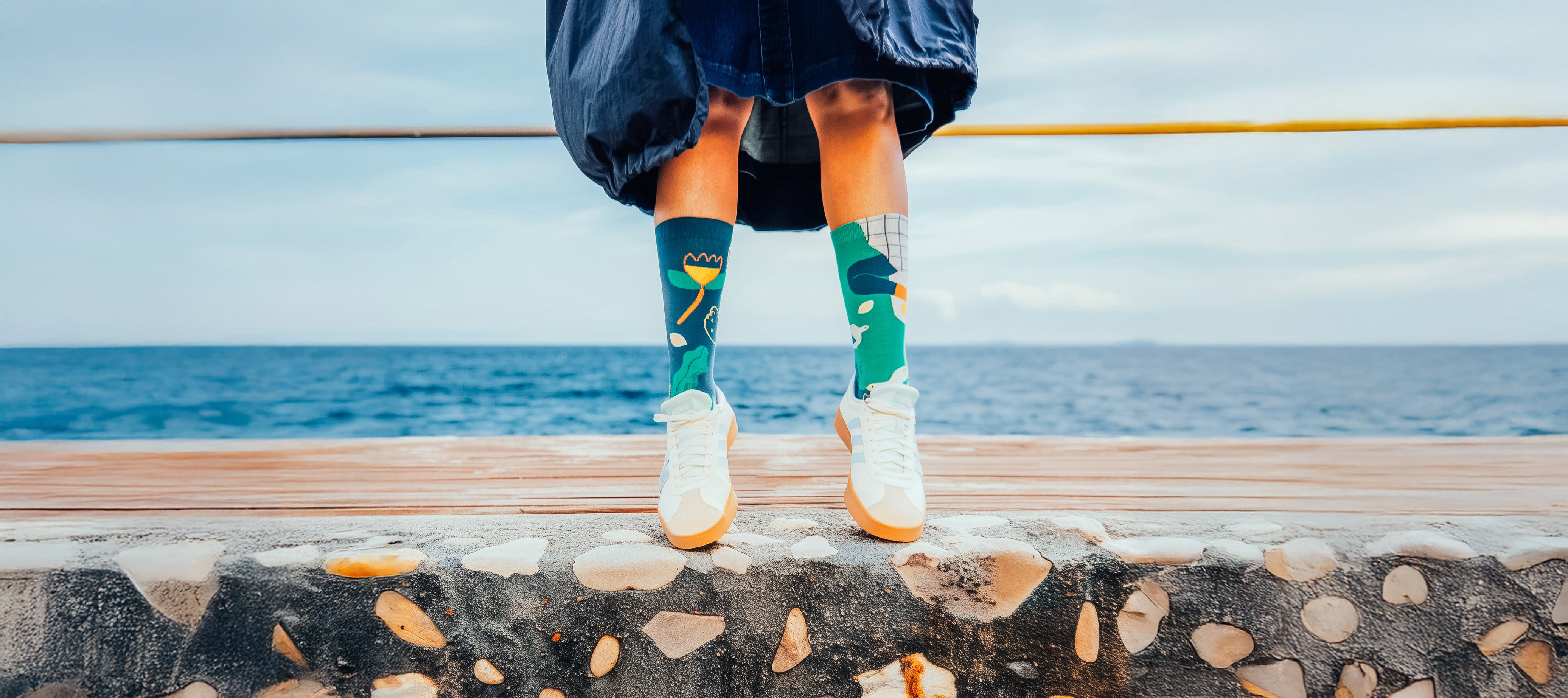 Person wearing colorful Limono Lemonheads socks and white sneakers standing on a stone ledge with water in the background