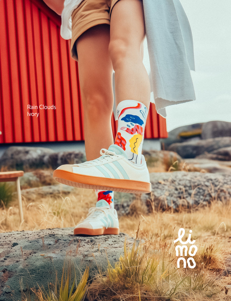 Limono Socks Rain Clouds collection. Illustrated socks on trend made in Portugal. Organic combed cotton. Person wearing colorful socks and white sneakers with a red building and natural landscape in the background