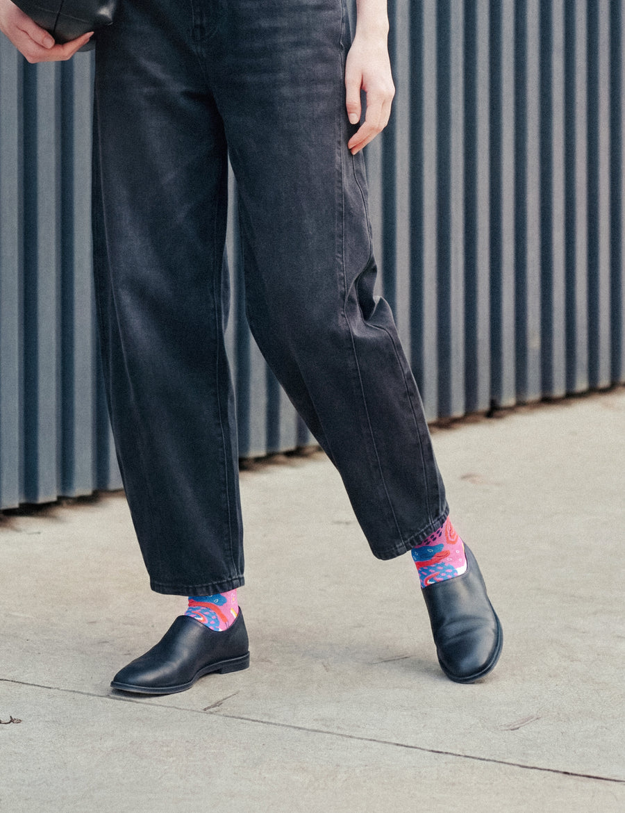 Woman in dark wide jeans and black flats wearing Limono Rain Clouds Magenta socks by a metal wall