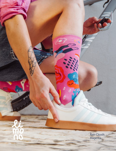 Woman in pink shirt putting on a Limono Rain Clouds Magenta sock with a white sneaker, outdoors
