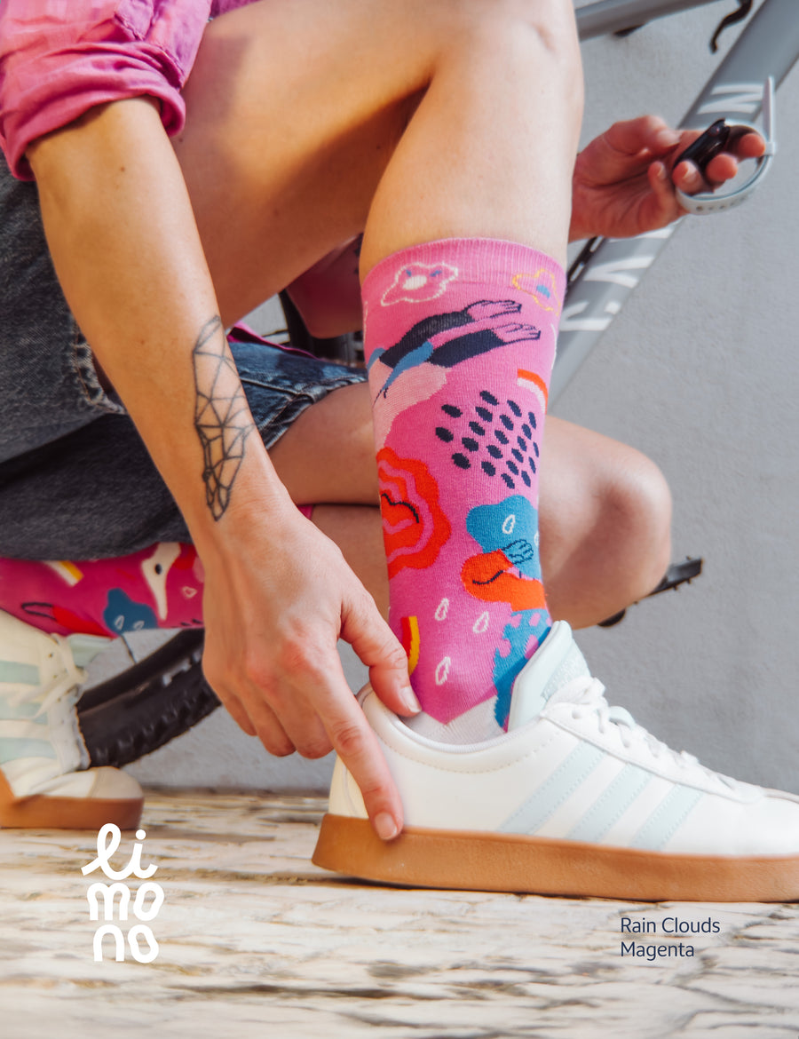 Woman in pink shirt putting on a Limono Rain Clouds Magenta sock with a white sneaker, outdoors