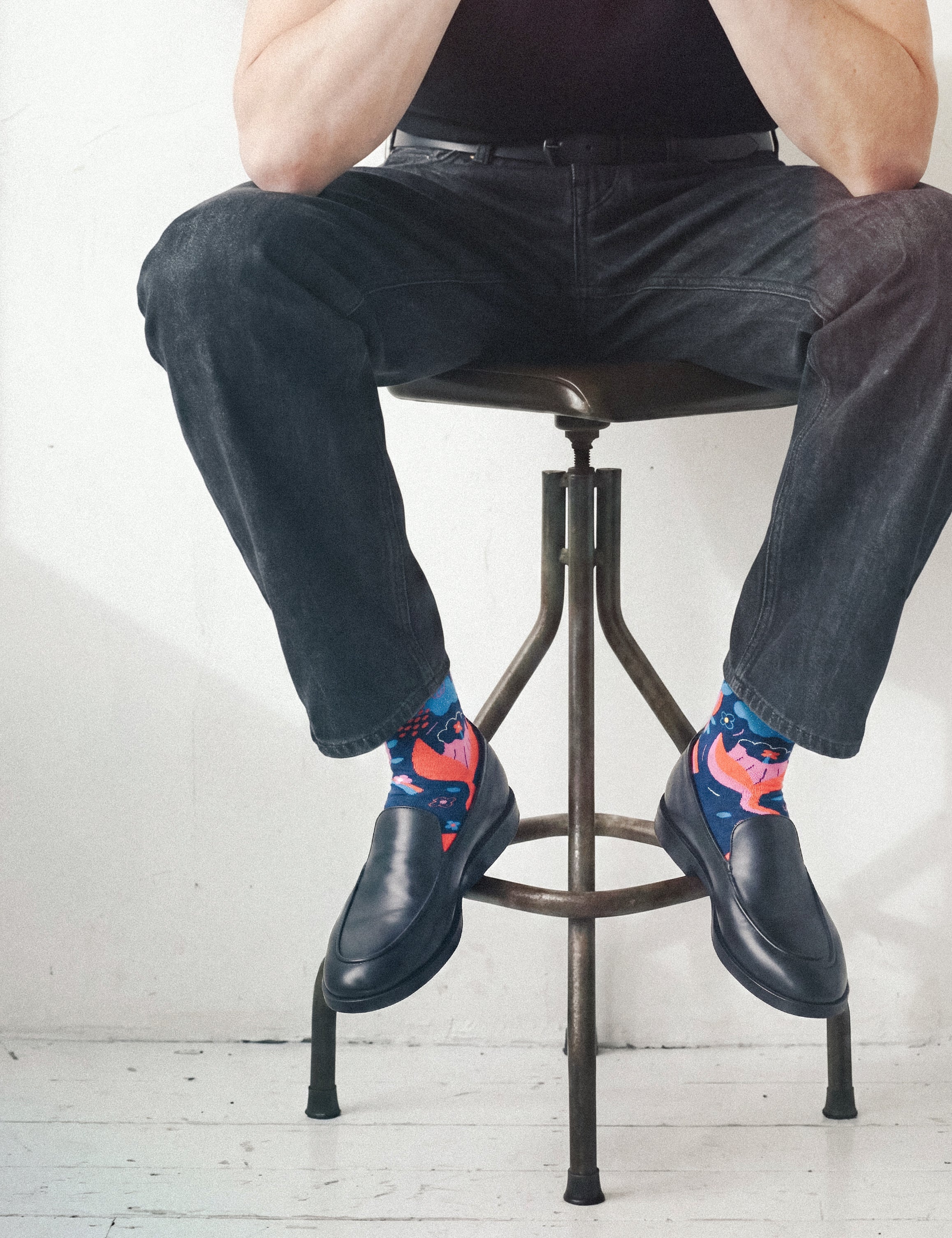 Man on a metal industrial bar stool in dark jeans and black loafers, wearing Limono Rain Clouds Black socks