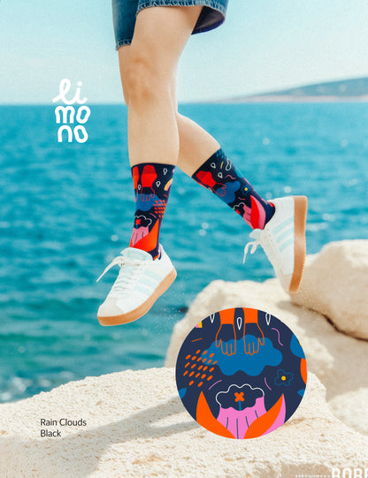 Limono Socks Rain Clouds collection. Illustrated socks on trend made in Portugal. Organic combed cotton. Person wearing colorful socks with a blue ocean background, featuring the brand 'liimo'.