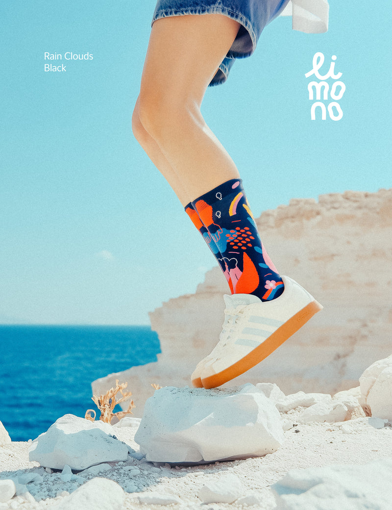 Limono Socks Rain Clouds collection. Illustrated socks on trend made in Portugal. Organic combed cotton. Person wearing colorful socks with a blue sky and ocean background, featuring the brand 'limo'.