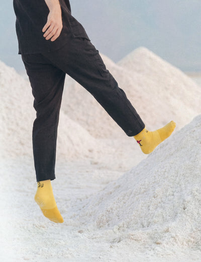 Limono Robots Day Yellow socks on a model mid-stride on salt mounds in a dark linen outfit