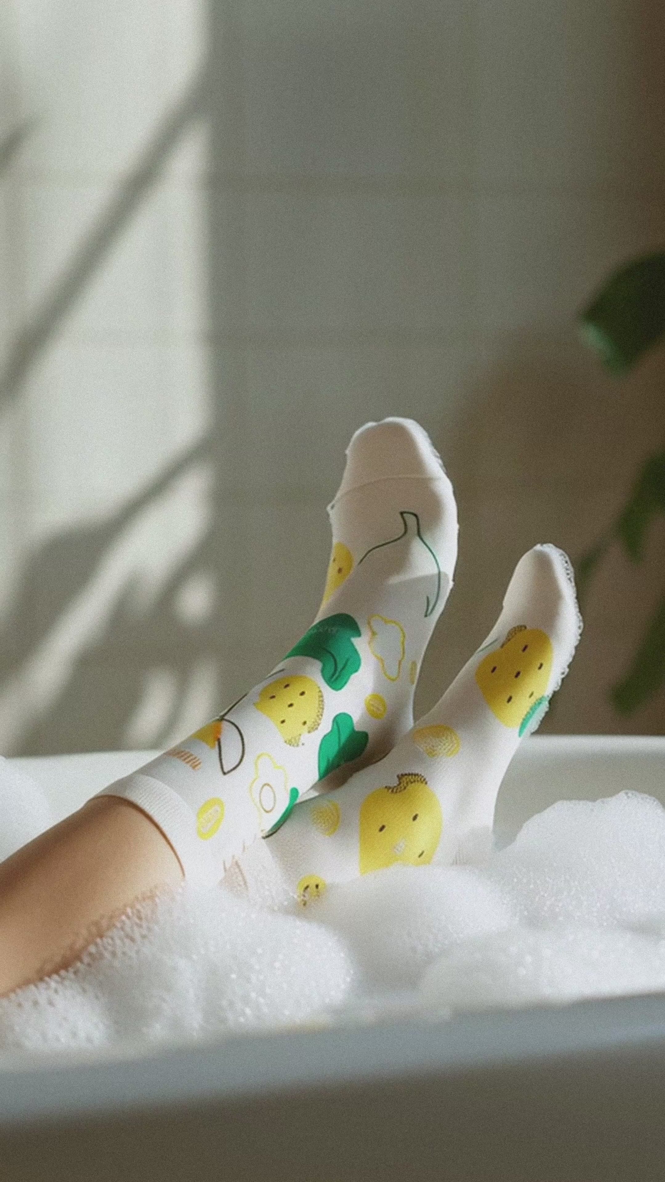 Load video: Limono Socks Lemonheads collection. Illustrated socks on trend made in Portugal. Organic combed cotton. Person wearing colorful Limono Lemonheads socks with a plant and text overlay on a light background laying in the bath foam.