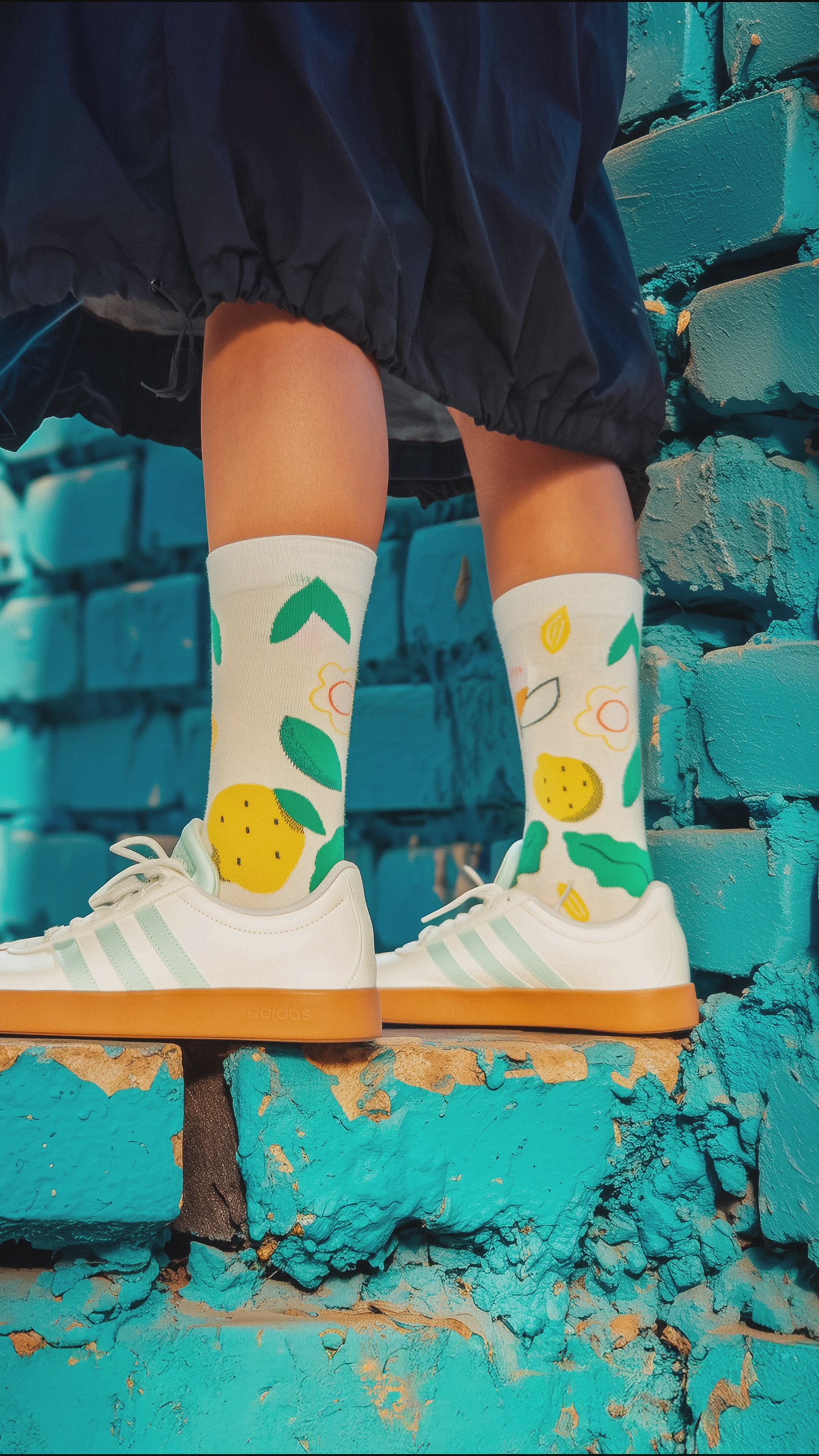 Load video: Person wearing colorful Limono Lemonheads socks and white Adidas sneakers against a blue brick wall.