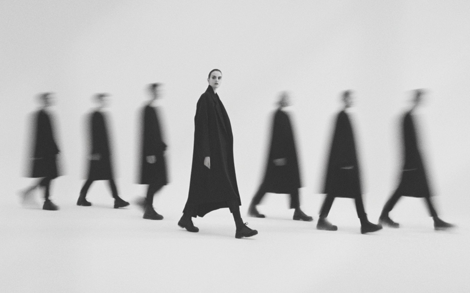 Black and white editorial of woman in long black coat with motion-blurred walking figures